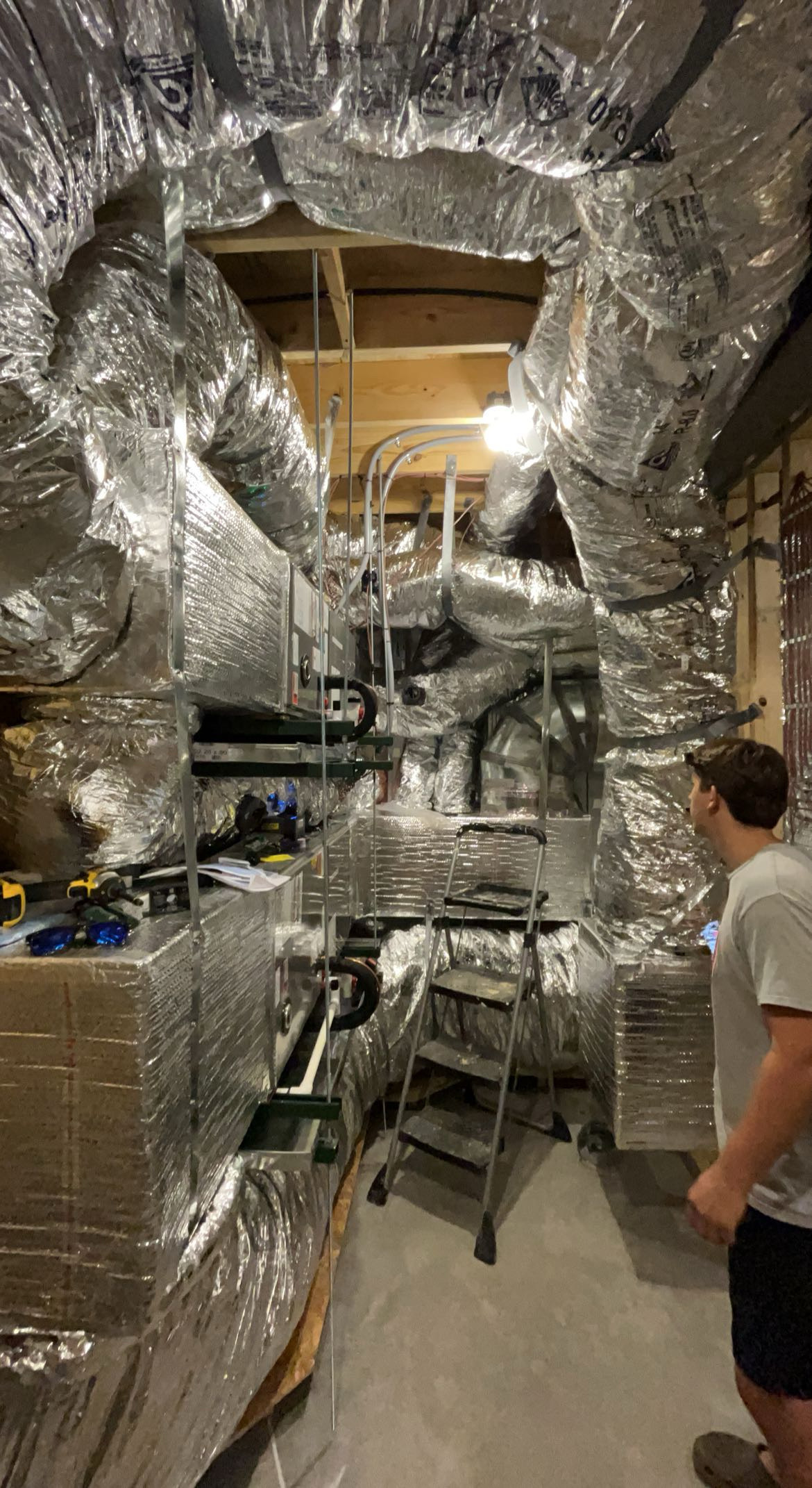 'n Persoon staan in 'n kamer vol HVAC-buiswerk bedek met reflektiewe isolasiemateriaal, met 'n leer en verskeie gereedskap sigbaar.