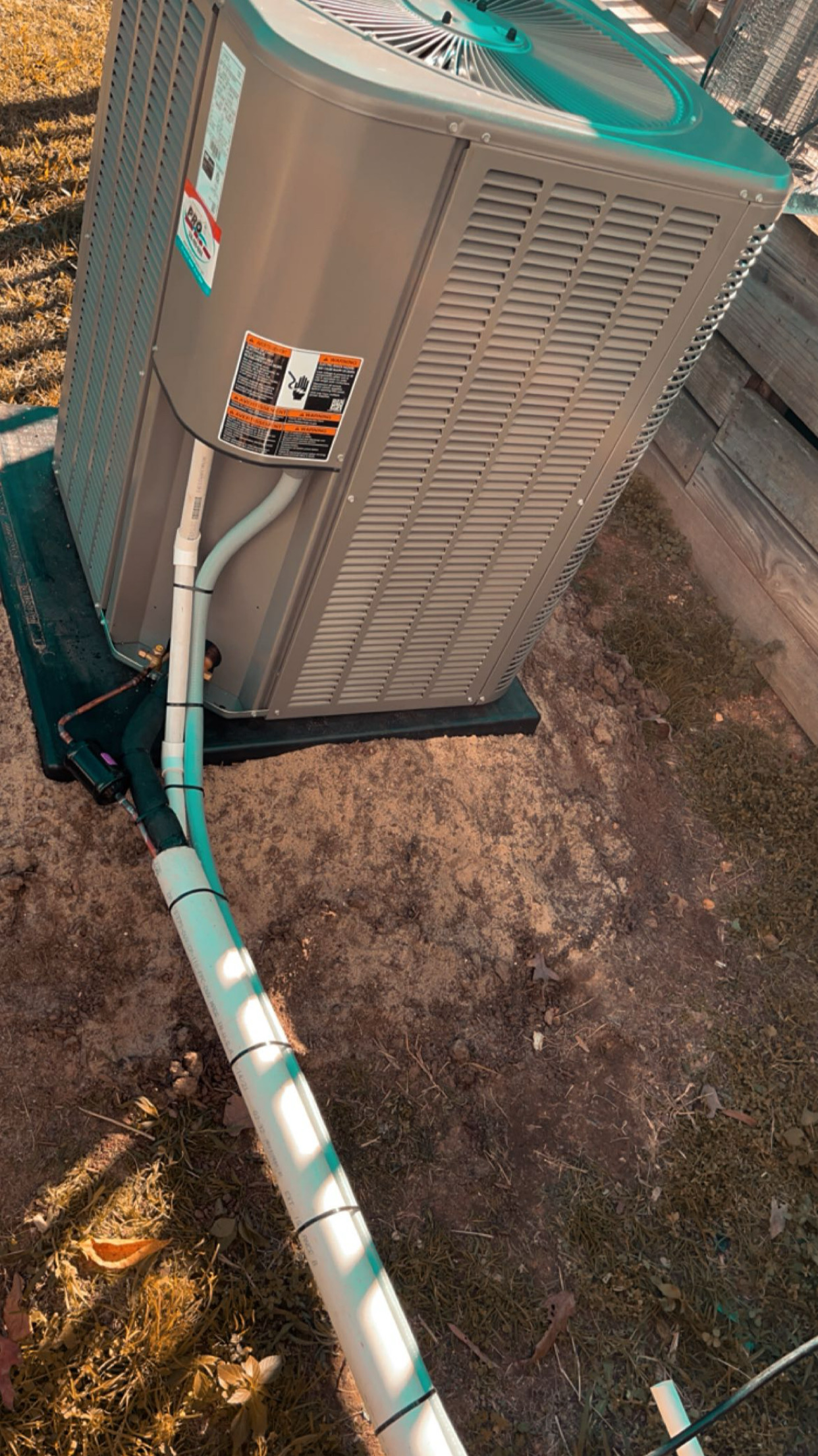 Outdoor air conditioning unit with connected pipes situated on a concrete pad next to a wooden fence, surrounded by grass and bare soil.