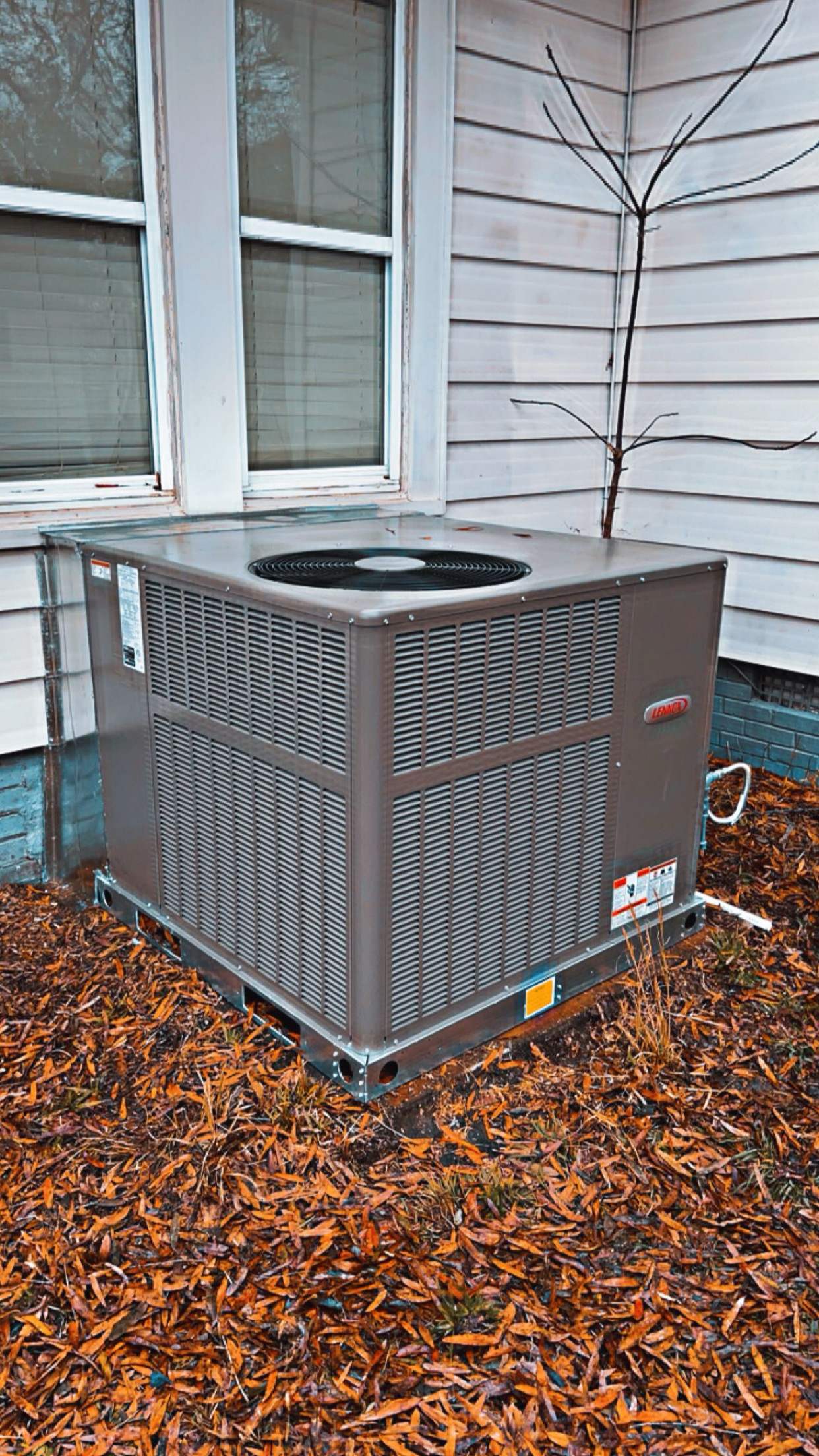 Outdoor air conditioning unit installed on the ground next to a house with white siding and windows; fallen leaves cover the surrounding ground.