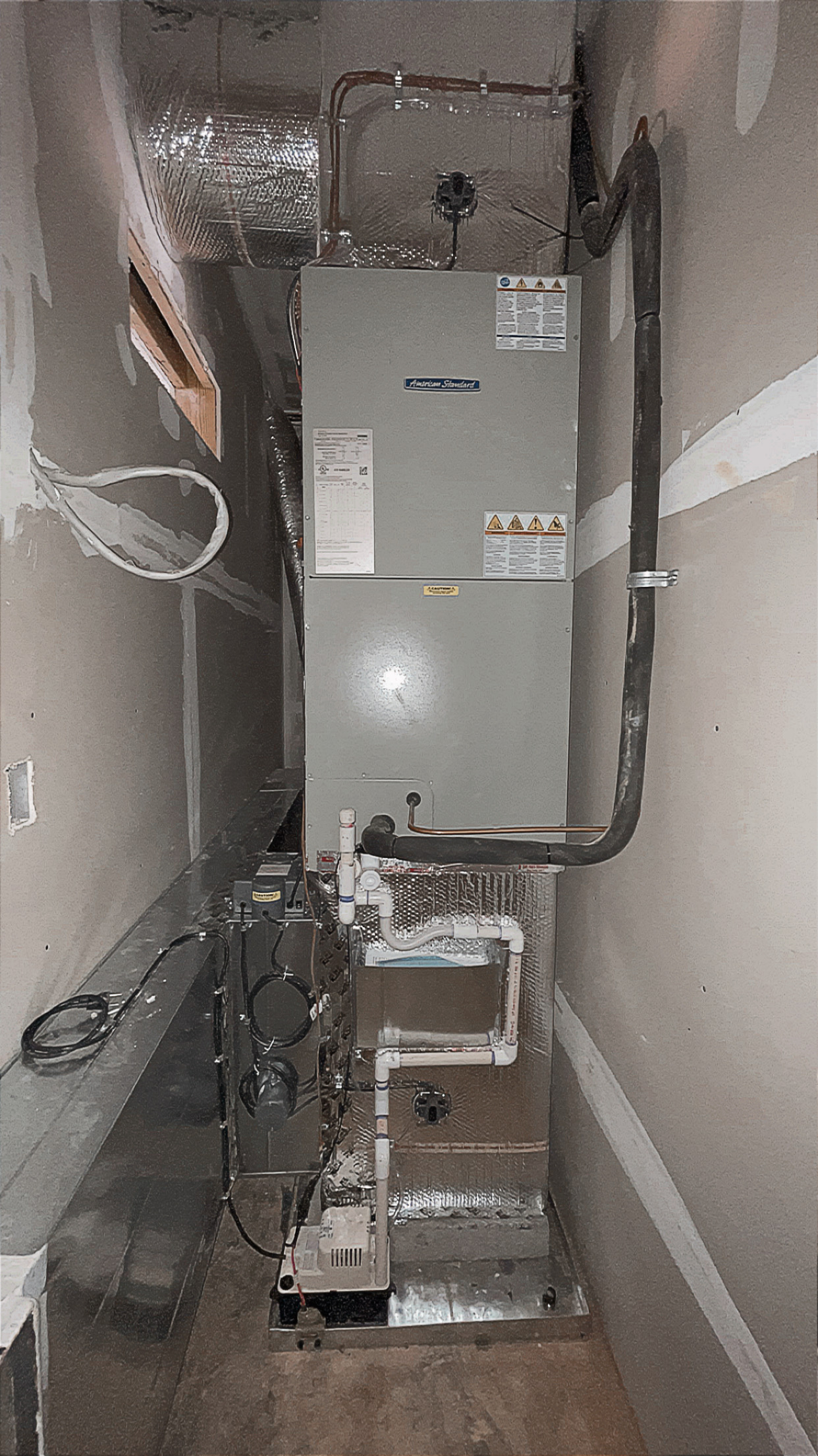 A central HVAC furnace unit with connected ductwork and pipes is installed in a narrow, unfinished utility room with exposed drywall.